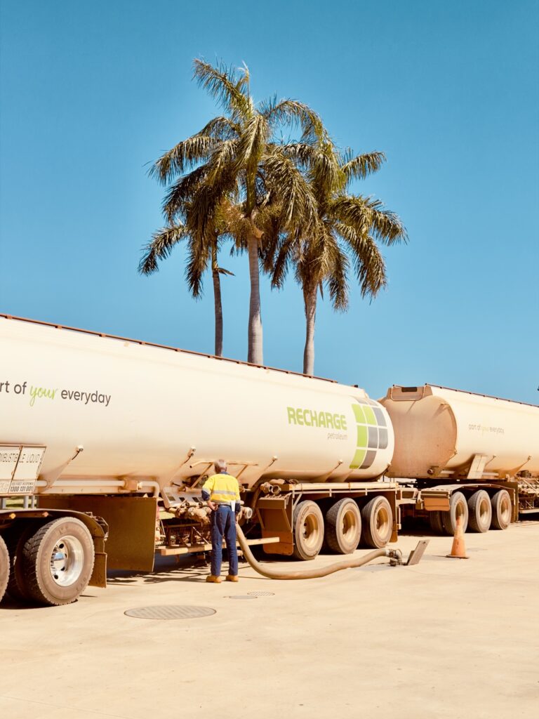 A road train tanker refuelling under tall palms