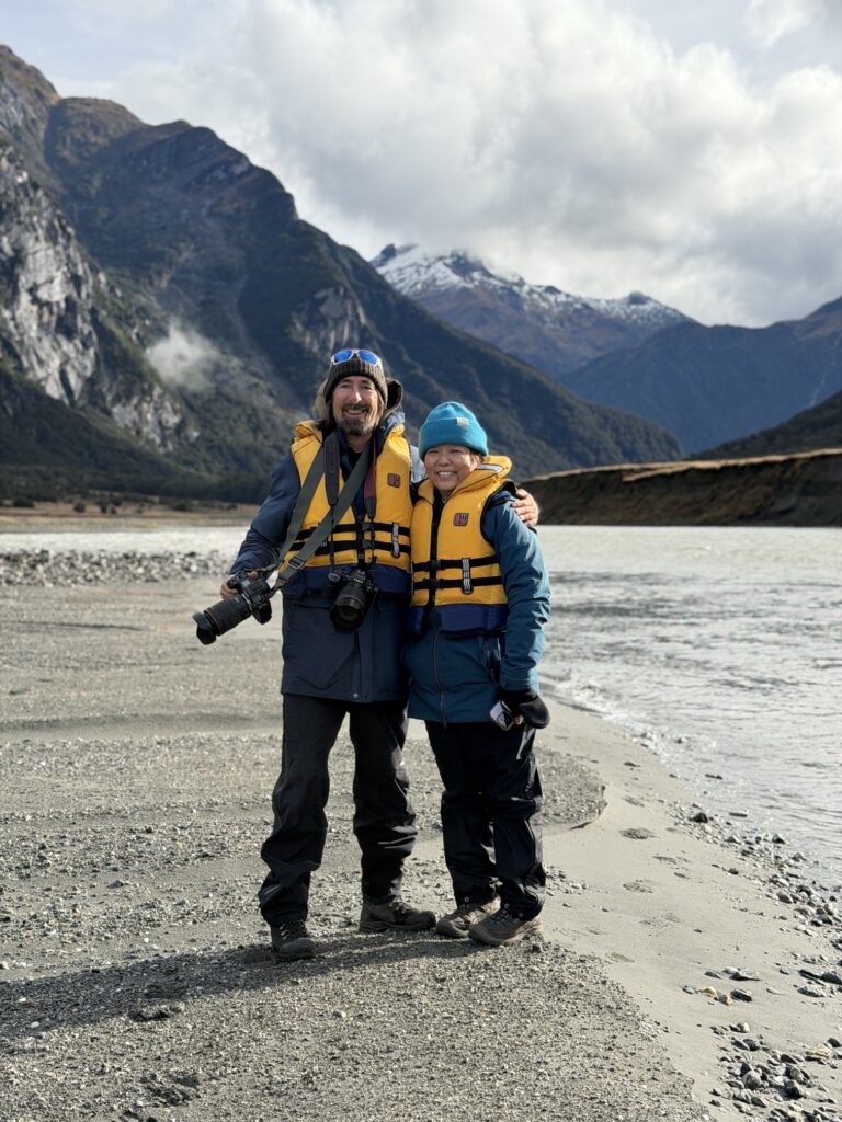 Two guests in life jackets on a river beach below the mountains